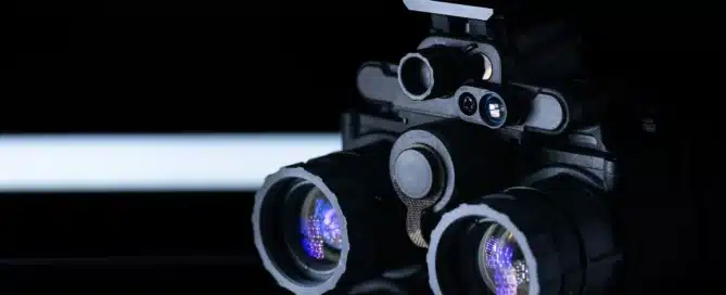 Dual-tube night vision goggles on a black reflective surface with violet lens reflections.