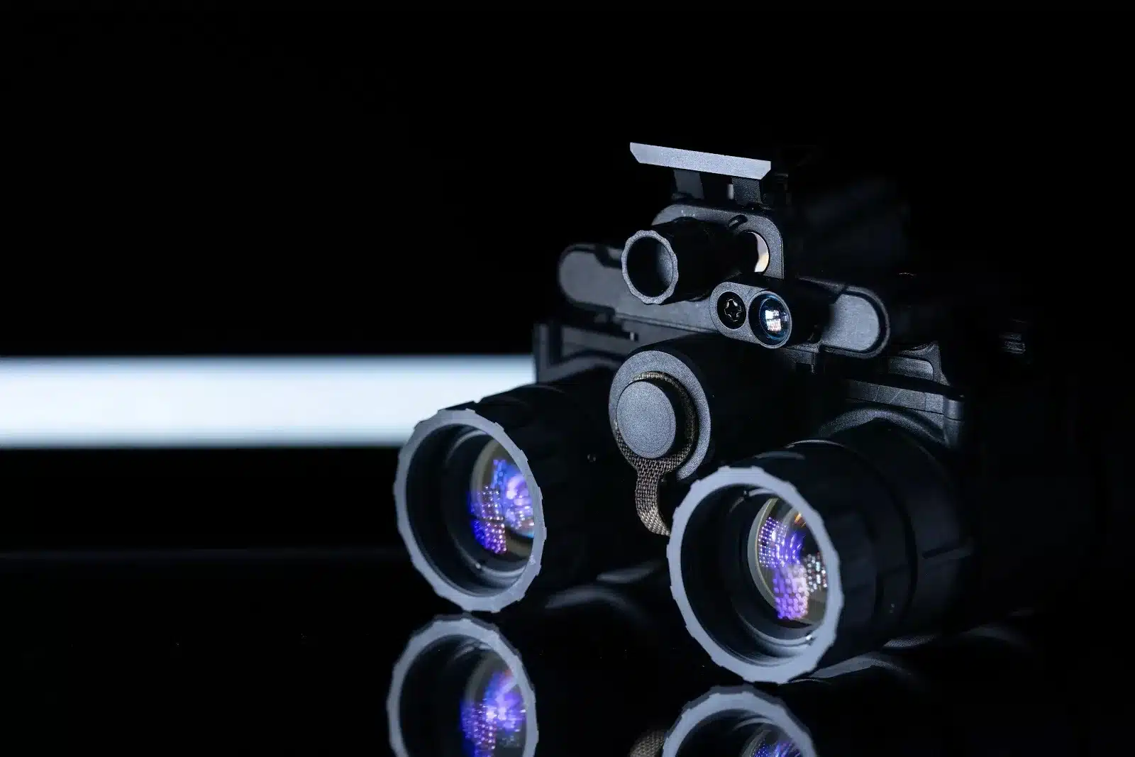 Dual-tube night vision goggles on a black reflective surface with violet lens reflections.